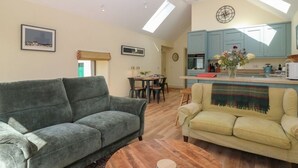 A spacious living area with a grey sofa, a yellow sofa, and a wooden coffee table. There is a dining table with chairs, a kitchen area with blue cabinets, and a skylight above the kitchen. - The Barn, Romantic Countryside Escape near St Andrews (Cupar)