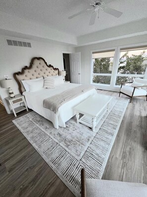 Quarto com uma cama grande, mesas de cabeceira, um banquinho, ventoinha no teto e vista pela janela ampla.