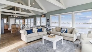 A spacious living room with a high ceiling, large windows, a kitchen area, and a view of the water. - Spacious Beach House | Decks & Pool + Beach Nearby (Bethany Beach)
