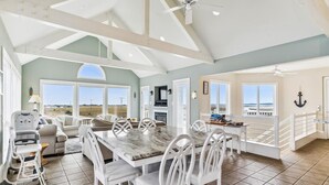 A spacious living area with a dining table, white chairs, a ceiling fan, and large windows offering a view of the ocean. - Spacious Beach House | Decks & Pool + Beach Nearby (Bethany Beach)