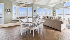 A spacious living area with a dining table, chairs, a sofa, and large windows offering a view of the water. - Spacious Beach House | Decks & Pool + Beach Nearby (Bethany Beach)