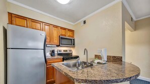 A compact kitchen with wooden cabinets, a stainless steel refrigerator, a microwave, and a granite countertop. - Sunset Cove – Tampa Sunsets from Your Balcony (Tampa)