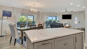 A modern kitchen with a large island, dining area with chairs, a flat-screen TV, and a ceiling fan. - Beachfront 3B Condo | Walk right onto the sand! (Indian Rocks Beach)
