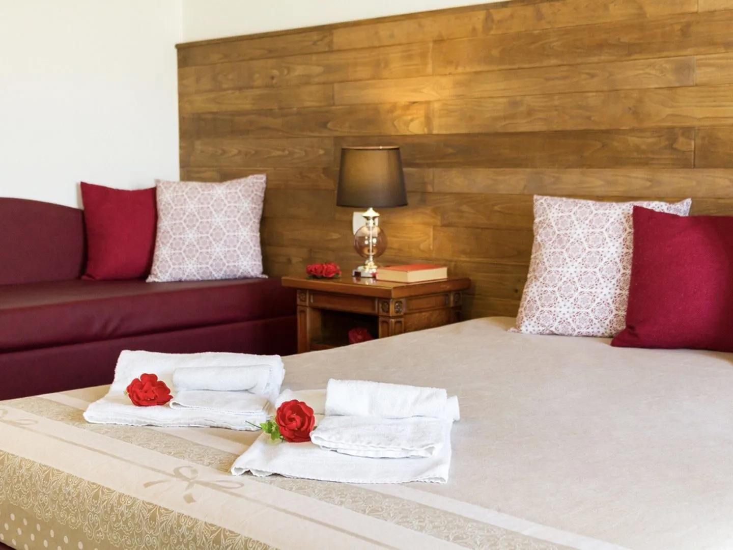 A hotel room with a wooden headboard, a bed with white linens, red pillows, a bedside table with a lamp, and a red rose on the bed.