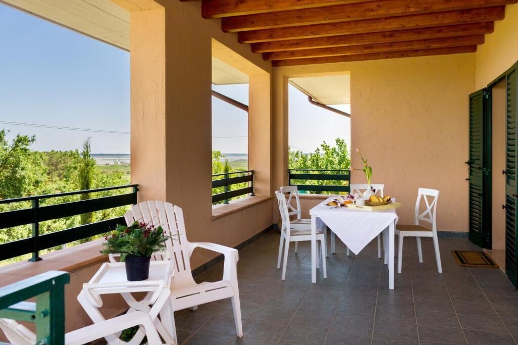 A balcony with white chairs and a table, a potted plant, and a view of greenery and a road.