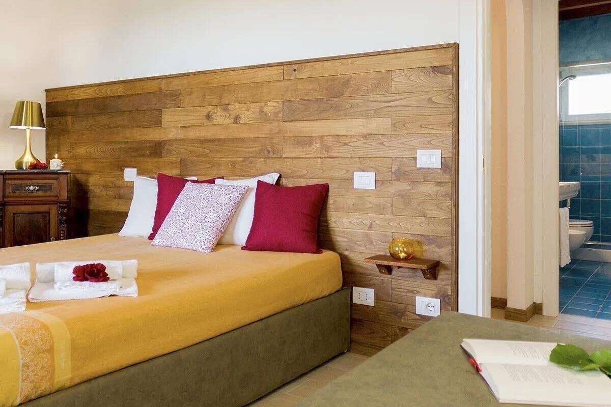Habitación de hotel con cabecera de madera, cama con ropa de cama amarilla, mesita de noche con lámpara y vista al baño.