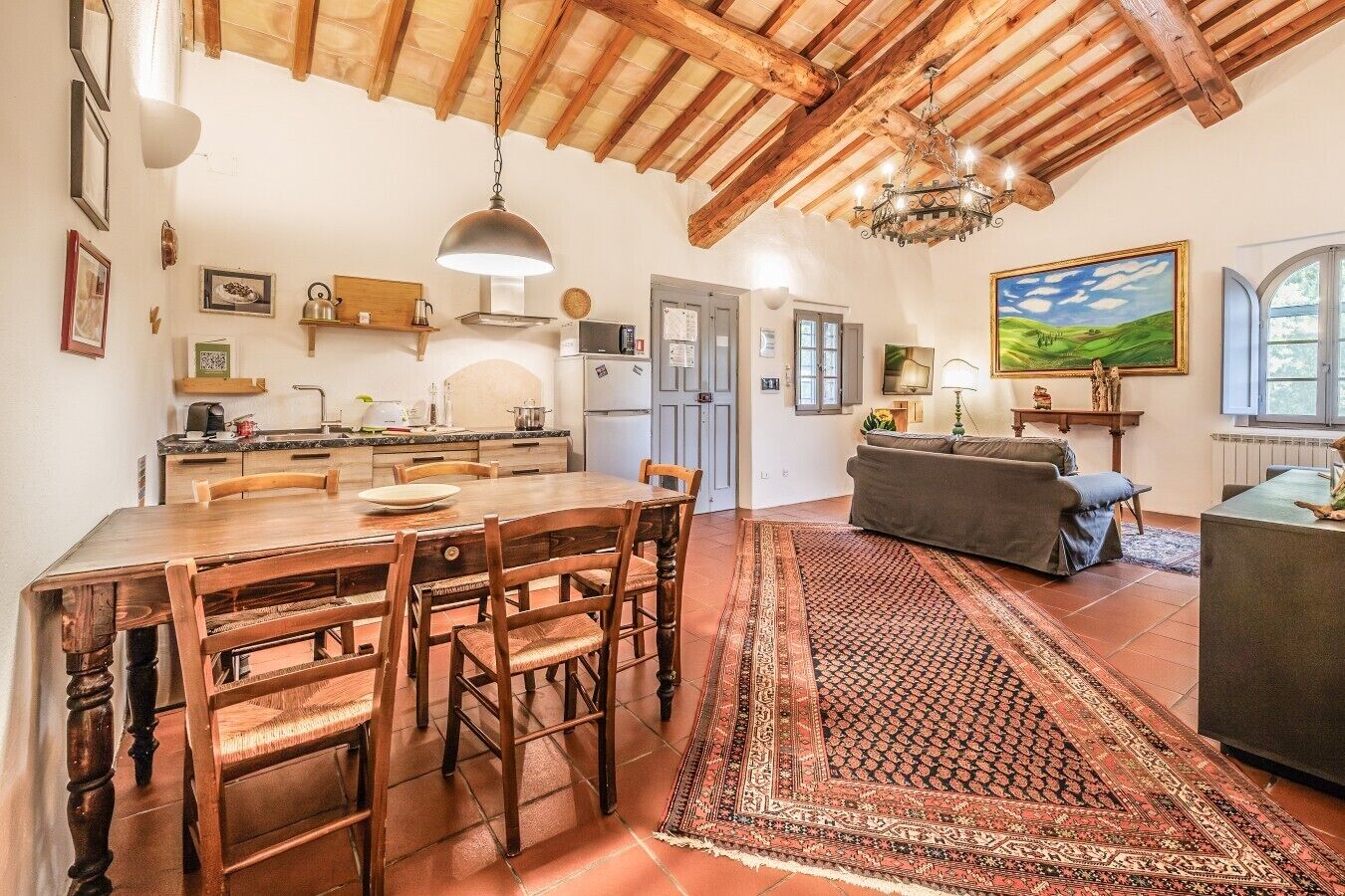 A spacious kitchen with wooden beams, a dining area with a large table and chairs, a living room with a sofa, and a painting on the wall.