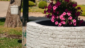 Un kiosque avec une base en briques et un toit en tuiles, entouré d’un jardin où poussent des fleurs roses, et un bâtiment blanc en arrière-plan.