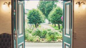 Une porte ouverte donnant sur un jardin où se trouvent un banc, un paillasson et un sol carrelé.