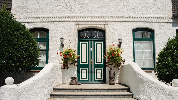 Un bâtiment blanc avec une porte et des fenêtres vertes, un escalier en pierre et des arrangements floraux de chaque côté.