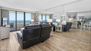 Moderni hotellihuone, jossa on suuri ikkuna, josta avautuu näkymä merelle, musta nahkasohva, työpöytä lampun kanssa sekä ruokailutila.