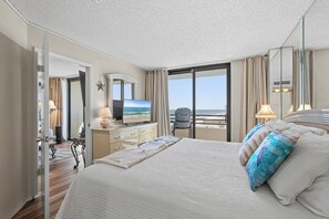 A hotel room with a large bed, a television, a desk with a lamp, and a view of the sea through the window. - 🌊Gorgeous Ocean Views | Oceanfront Complex | Sand Dollar 1105🌊 (Daytona Beach Shores)