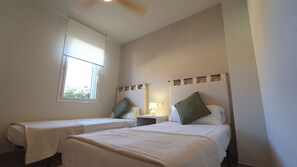 Ein Zimmer mit zwei Betten, einem Fenster, einem Nachttisch mit Lampe und einem Deckenventilator.