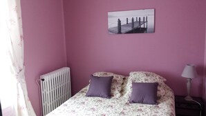 A bedroom with a pink wall, a bed with floral bedding and two pillows, a radiator, a picture of a pier, and a lamp on a bedside table. - Les Lauriers Roses (Bollène)