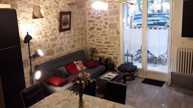 A cozy living room with a stone wall, a sofa with red cushions, a dining table with a floral tablecloth, and a chair. - Les Lauriers Roses (Bollène)