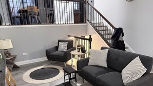 A modern living room with a grey sofa, armchair, and ottoman, a round coffee table, and a staircase with a railing. - Modern Spacious Retreat with Fast Wi-Fi, Yard & Workspace - 4BR and 2 Full Bath (Waukegan)