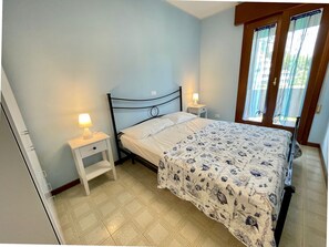 A bedroom with a bed, two bedside tables, a nightstand, a window with curtains, and a door. - Spacious Beachside Flat With Terrace (Caorle)