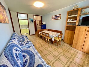 A compact living space with a sofa, dining table, chairs, a kitchen area, and a cabinet with a television. - Spacious Beachside Flat With Terrace (Caorle)
