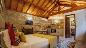 A bedroom with a stone wall, wooden ceiling beams, a bed with pillows, a TV, a microwave, and a small kitchenette. - Refuge of Plenitude (Canelas)