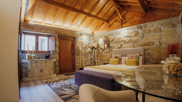 A cozy room with a stone wall, wooden ceiling beams, a glass table, and a bed with colorful pillows. - Refuge of Plenitude (Canelas)