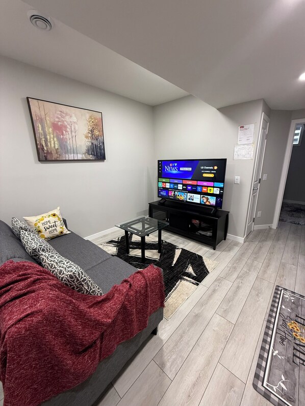 A modern living room with a grey sofa, a red blanket, a glass coffee table, and a flat-screen TV displaying news channels.