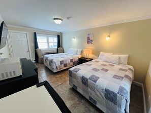 A hotel room with two beds, a desk, a chair, a lamp, and a painting on the wall. - Dolan's Inn & Suites (Pictou)