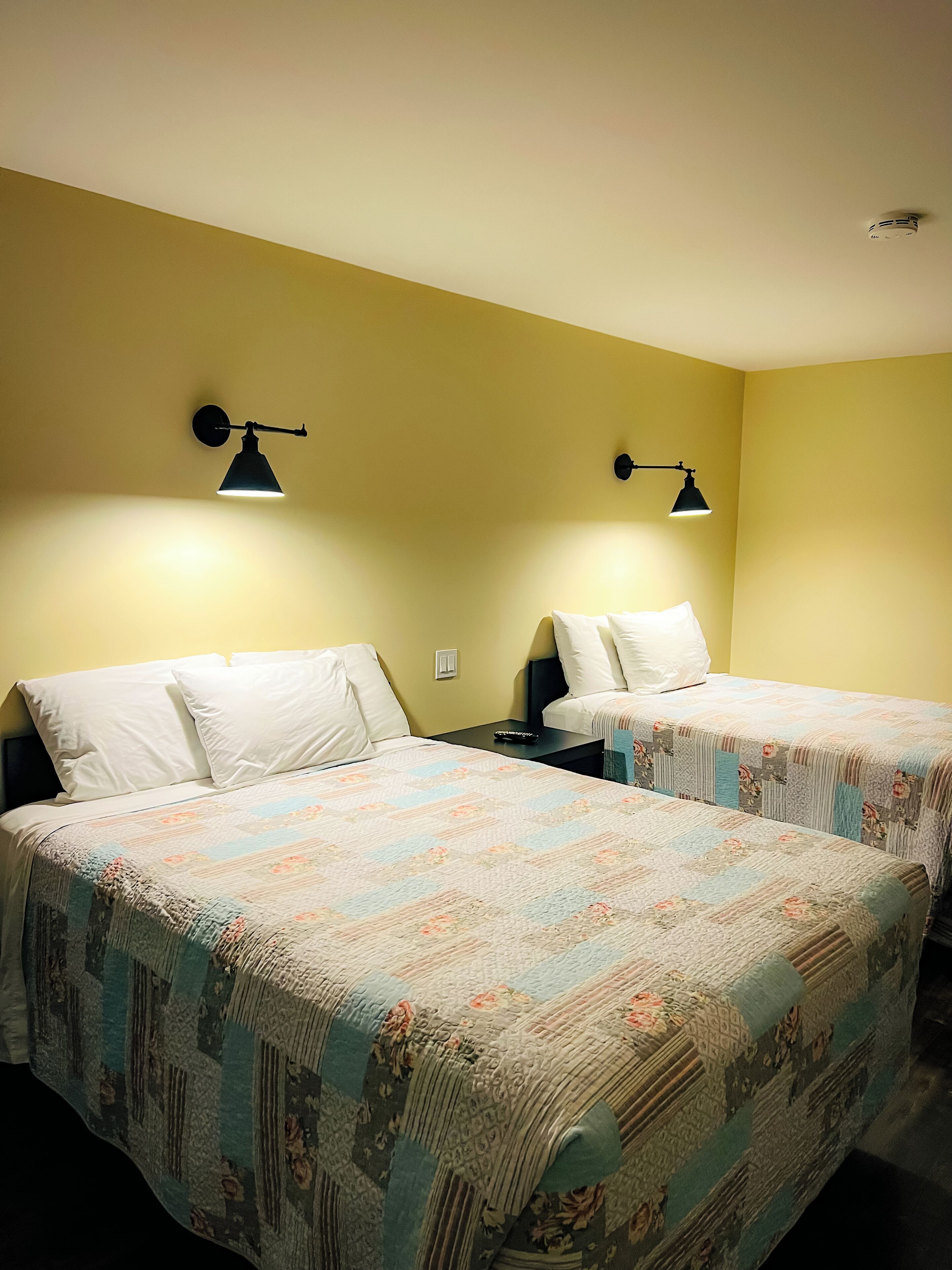 A room with two beds, each with a patterned bedspread and white pillows. There are two wall-mounted lamps providing light.