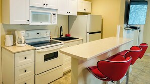 A compact kitchen with white cabinets, a microwave, a stove, a refrigerator, and a bar area with red chairs.