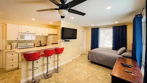 A compact hotel room with a kitchenette, a bed, a TV, and a balcony with curtains.