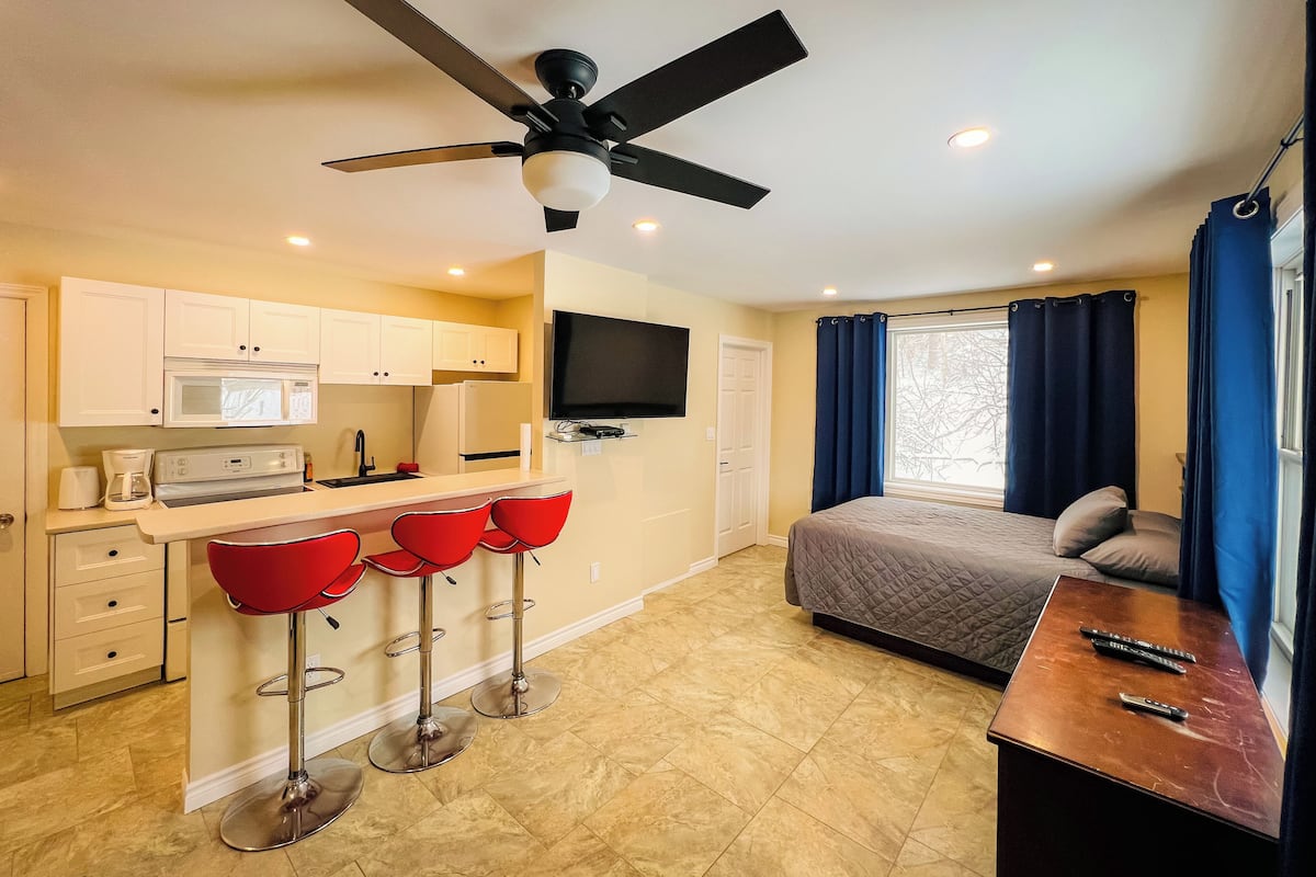 A compact hotel room with a kitchenette, a bed, a TV, and a balcony with curtains.