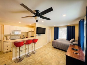 A compact hotel room with a kitchenette, a bed, a TV, and a balcony with curtains.