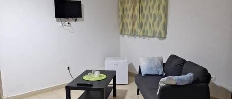 A room with a bed, a black sofa, a small table with a green placemat, a white air conditioner, and a TV mounted on the wall.