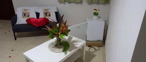 A room with a bed, a small white table with a plant, a white cabinet, and a red pillow on the bed.