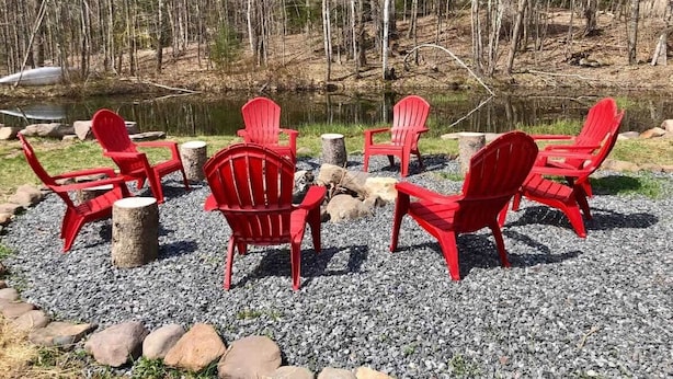 Cadeiras Adirondack vermelhas dispostas em círculo ao redor de uma lareira, sobre um piso de cascalho, com uma floresta ao fundo.