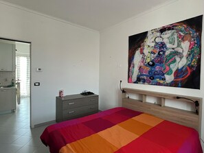 Ein Schlafzimmer mit Bett, einem Schminktisch, einem Gemälde an der Wand und einem durch eine geöffnete Tür erreichbaren Badezimmer.