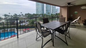 Ein Balkon mit Tisch und Stühlen, Deckenventilator und Blick auf das Meer und hohe Gebäude.