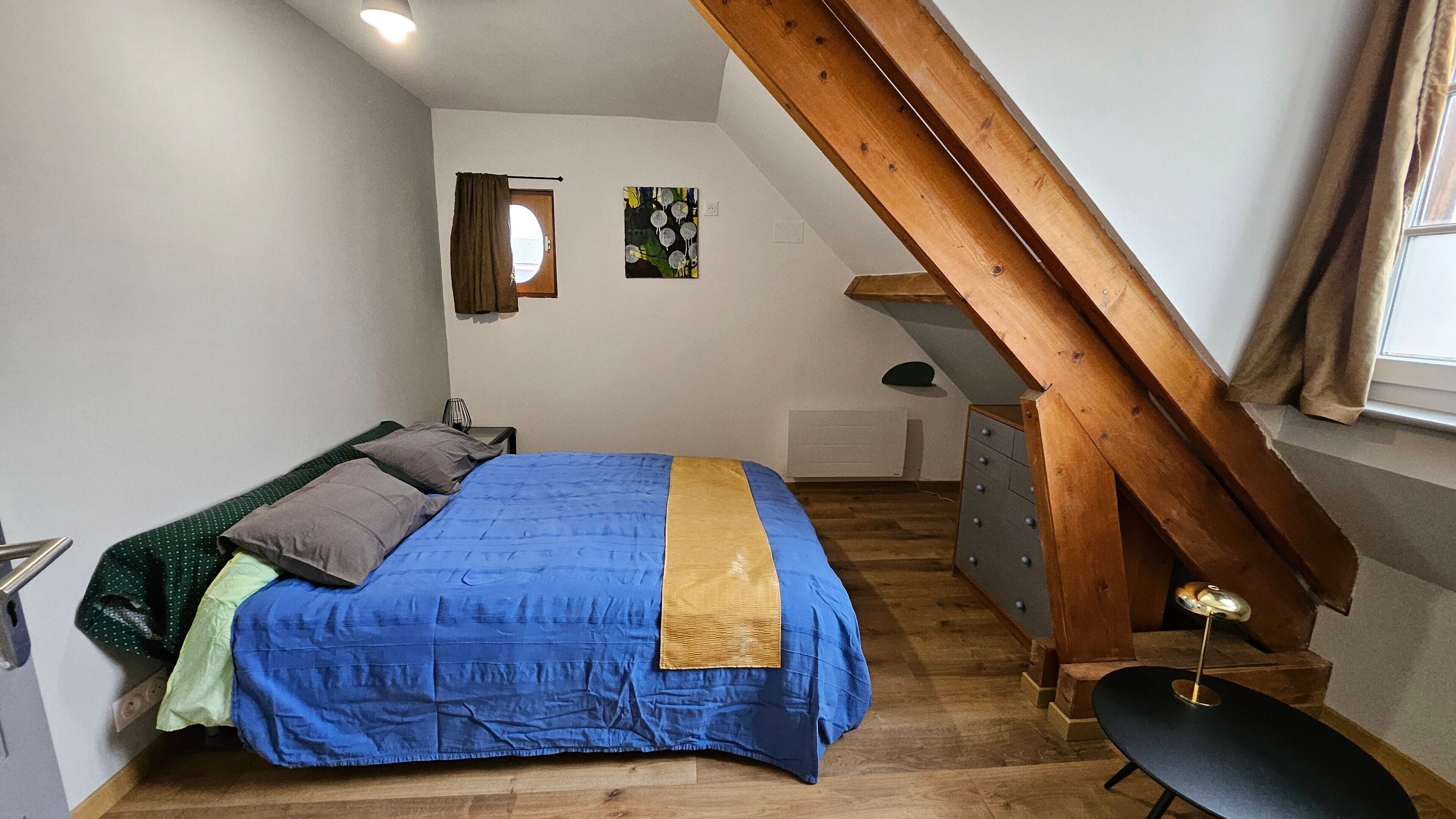 A bedroom with a bed, a dresser, a chair, a window with curtains, a painting, and a sloped ceiling.