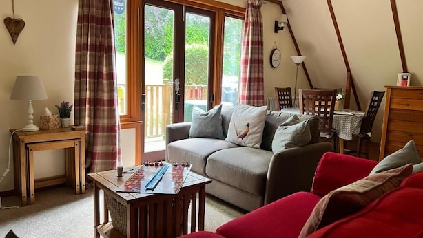 Un salon chaleureux avec un canapé, une table sur laquelle est posé un jeu de société, une lampe et une vue dégagée sur l’extérieur grâce à une grande fenêtre.