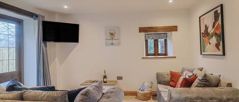 A living room with two grey sofas, a small table with a bottle and glass, a TV mounted on the wall, a window with curtains, and a framed picture.