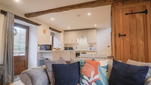 A cozy living room with a sofa, pillows, and a wooden ceiling beam. A kitchen is visible in the background with white cabinets and a microwave. - Rose Cottage (Wellington)
