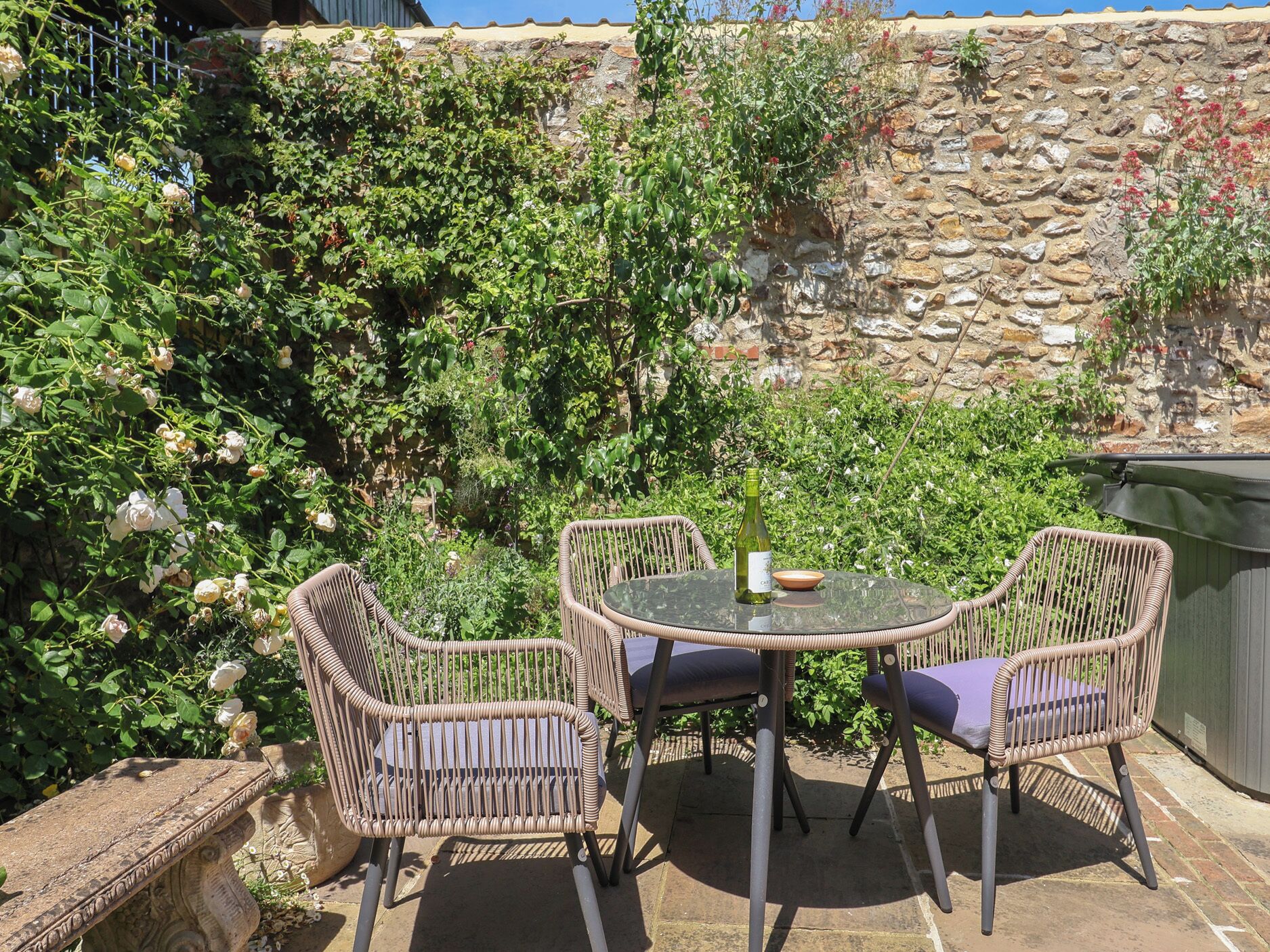 An outdoor patio with wicker chairs and a glass table, surrounded by lush greenery and a stone wall.