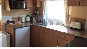 A compact kitchen with wooden cabinets, a microwave, a refrigerator, a toaster, and a window with floral curtains.