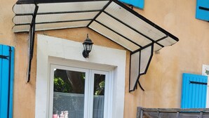 A house entrance with a white door, blue shutters, a black awning, and a wooden fence.
