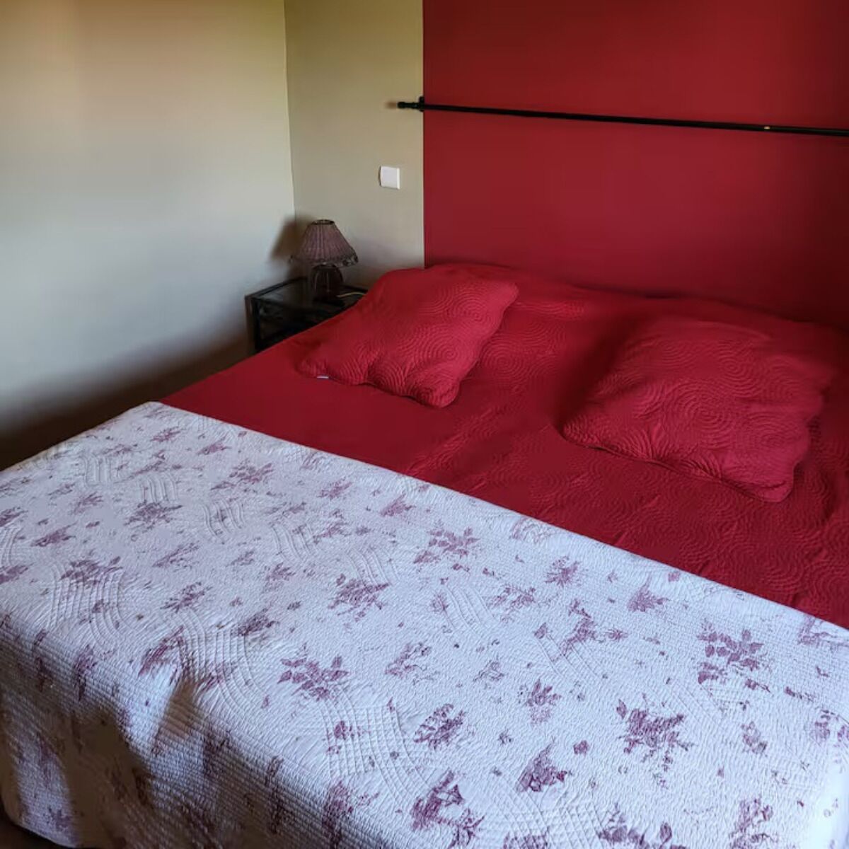 Een slaapkamer met een rode sprei, een wit bloemenbed en twee rode kussens.