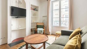 Ein Wohnzimmer mit einer Couch, einem runden Holztisch, einem Bücherregal, einem an der Wand montierten Fernseher und einem Fenster mit Vorhängen.