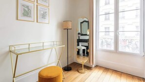 Ein Raum mit einem Glastischchen, einem runden Hocker, einer Stehlampe und einem Spiegel, der einen Stuhl und einen Tisch reflektiert.