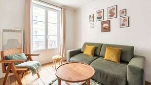 Ein Wohnzimmer mit einem grünen Sofa, einem Holzstuhl und einem runden Tisch. Sechs gerahmte Kunstwerke hängen an der Wand.