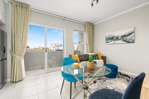 A modern living room with a glass dining table, blue chairs, a sofa with cushions, a cactus plant, and a large abstract painting on the wall. - The Orion 16 (Cape Town)