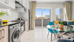 Une cuisine moderne équipée d’un lave-linge, un coin repas avec une table en verre et un balcon avec des rideaux.