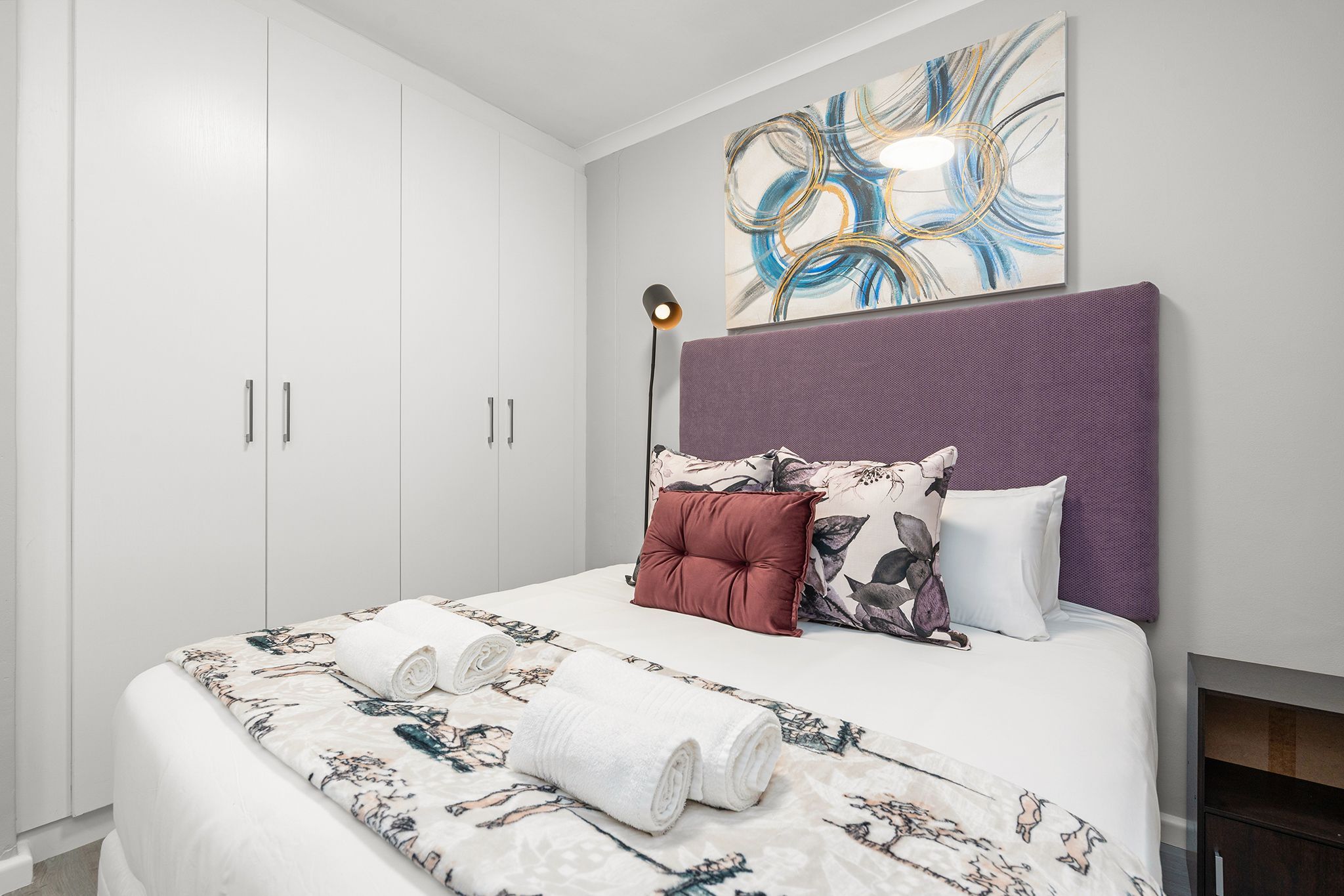 A bedroom with a large bed, a headboard, a wall art piece, and a floor lamp.